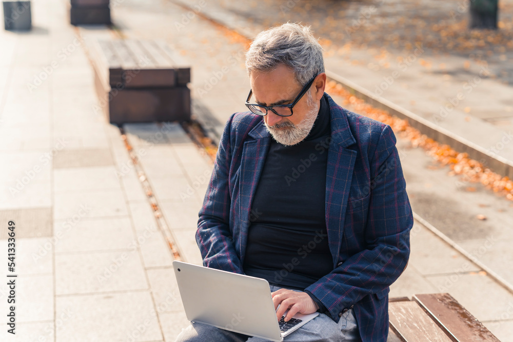 Modern stylish bearded grey-haired mature man entrepreneur sitting on a bench in the park with fall foliage and working on his laptop. High quality photo Modern stylish bearded grey-haired mature man entrepreneur sitting on a bench in the park with fall foliage and working on his laptop. High quality photo