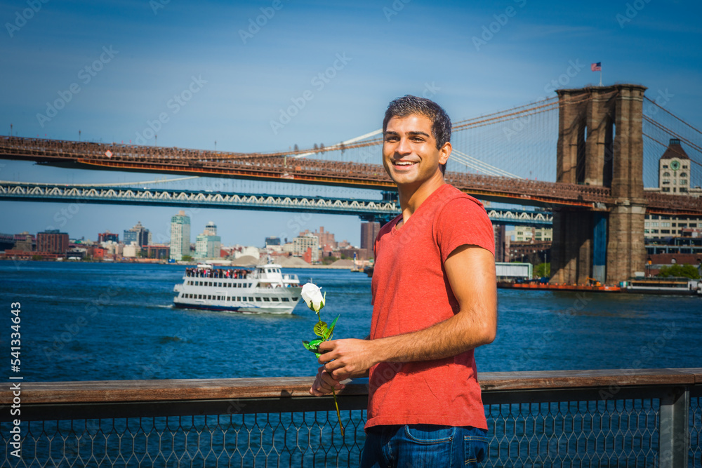 Young East Indian American man wearing red V neck T shirt, jeans, standing by river, holding white rose, smiling, waiting for you. Manhattan, Brooklyn bridges, boat on background. .