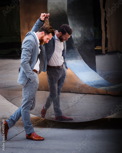 Frustration. American Business Man with beard working in New York City, wearing cadet blue suit, brown leather shoes, standing by metal mirror wall, looking down, sad, into deeply thinking..