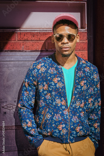 Obraz na płótnie Young African American Man wearing blue flower patterned, collarless jacket, green V neck shirt, yellow brown pants, pink cap, sunglasses, standing against wall under sun on street in New York City.. w salonie