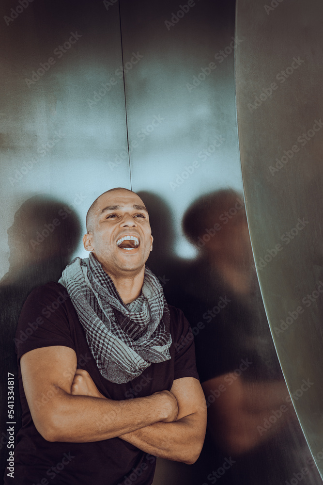 Young man with shaved head, wearing black T shirt, long patterned scarf around neck, crossing arms, standing in corner, opening mouth, shouting. .