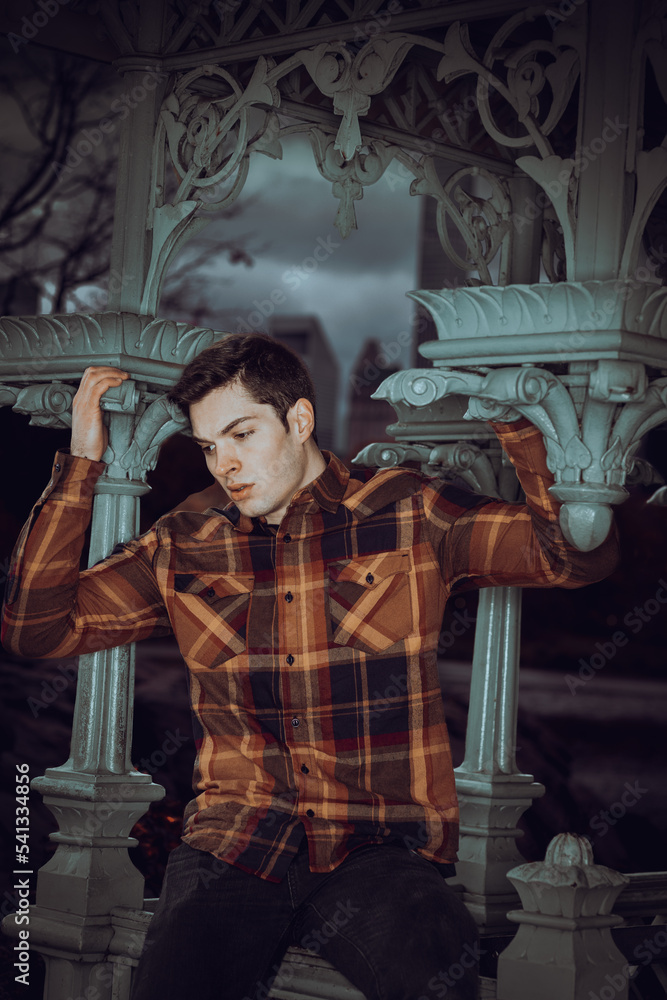 Young man wearing brown patterned long sleeve shirt, black jeans, siting on railing of pavilion in New York City in cloudy day, looking down sad, thinking.