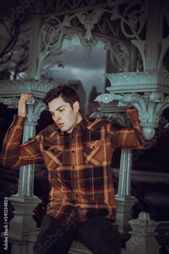 Obraz na płótnie Young man wearing brown patterned long sleeve shirt, black jeans, siting on railing of pavilion in New York City in cloudy day, looking down sad, thinking. w salonie