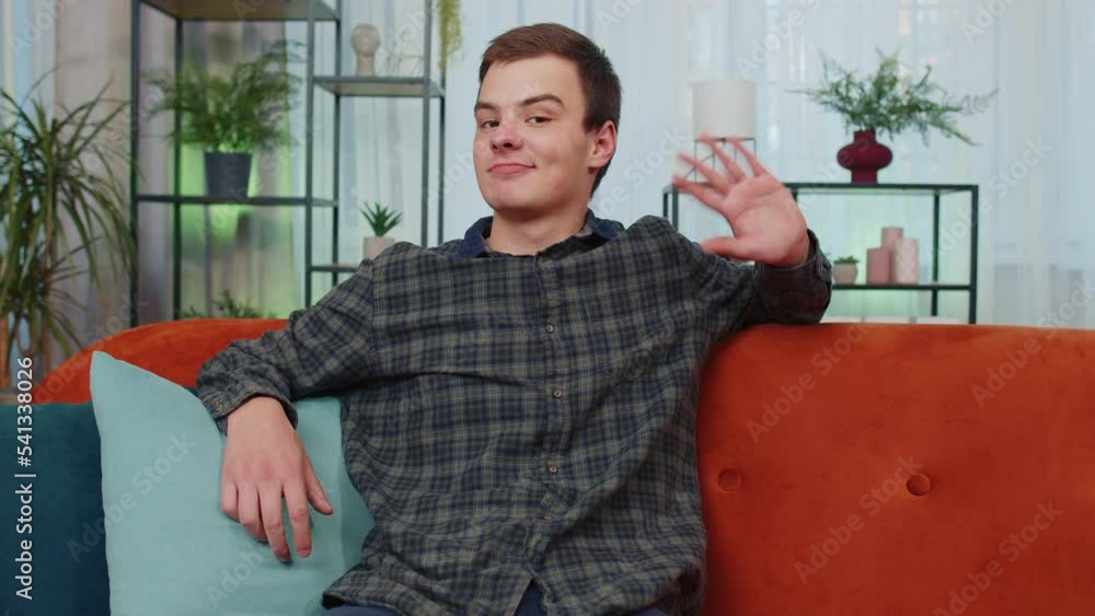 Teenager man student smiling friendly at camera and waving hands gesturing hello, hi, greeting or goodbye, welcoming with hospitable expression. Young guy boy sitting on orange couch at home room Teenager man student smiling friendly at camera and waving hands gesturing hello, hi, greeting or goodbye, welcoming with hospitable expression. Young guy boy sitting on orange couch at home room