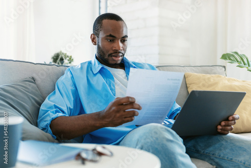 Paperwork concept. Portrait of black man working at home, reading paper or financial document, sitting on sofa