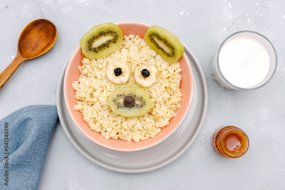 Funny cute kids childrens baby's healthy breakfast lunch oatmeal porridge in bowl look like bear,mouse face decorated with kiwi, banana, dried berry fruits,milk,honey. dessert food art on gray table