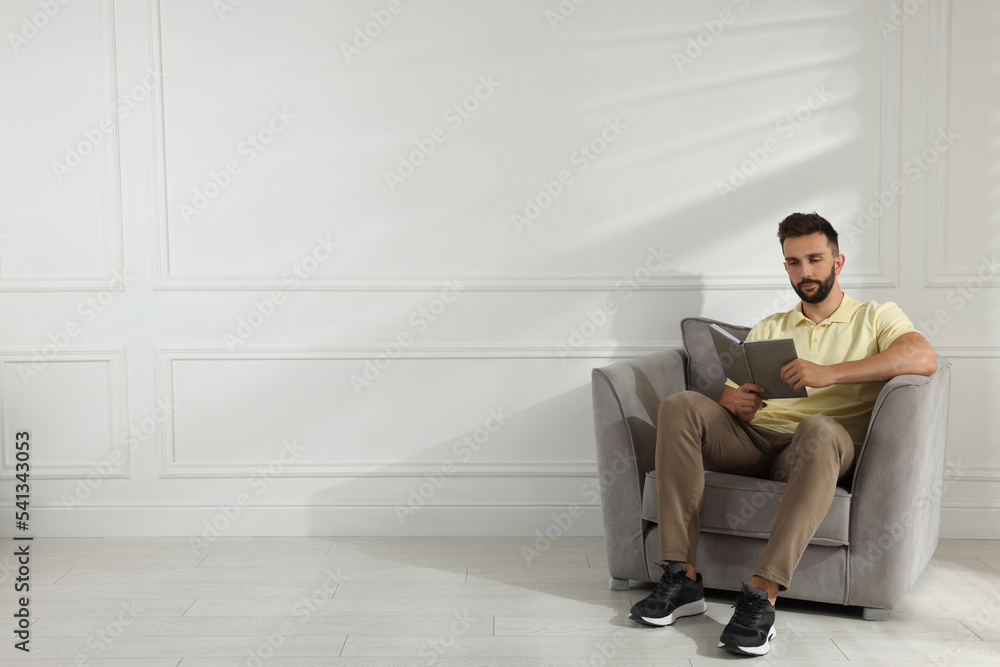 Man reading book in armchair near white wall, space for text Man reading book in armchair near white wall, space for text
