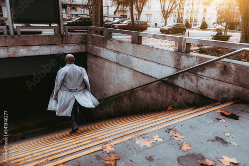 View from behind of a mature elegant bald black businessman in a grey overcoat descending to a subway station, the floors of the coat flutter in the wind; a hairless African guy going down to a metro