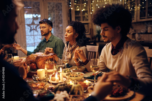 Young black woman and her friends praying while having dinner together on Thanksgiving.