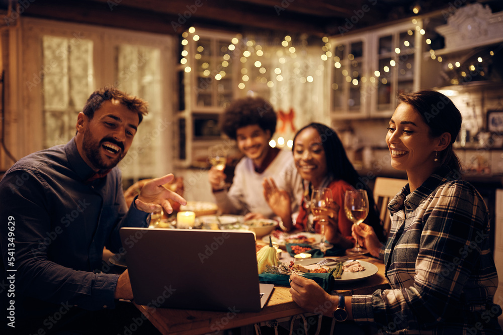 Happy friends making video call via laptop while having dinner at dining table on Thanksgiving. Happy friends making video call via laptop while having dinner at dining table on Thanksgiving.