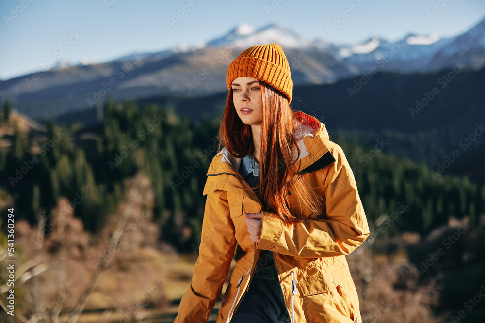 Woman hiking in the mountains in the fall with a smile with teeth and happiness in a yellow cape with red hair full-length stands against the backdrop of trees and mountains in the sunset light