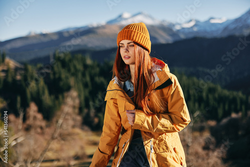 Woman hiking in the mountains in the fall with a smile with teeth and happiness in a yellow cape with red hair full-length stands against the backdrop of trees and mountains in the sunset light © SHOTPRIME STUDIO