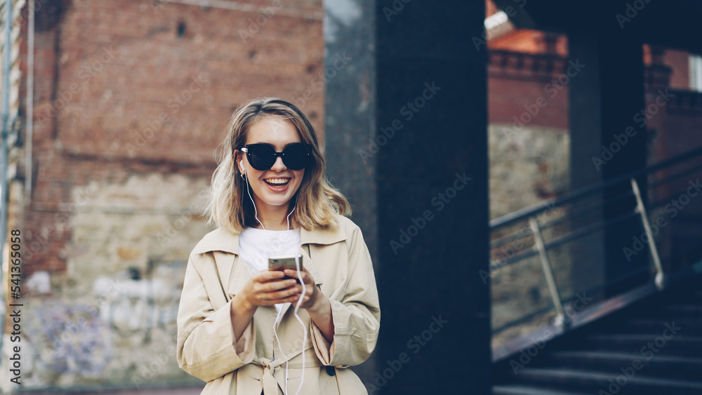 Fototapeta premium Pretty girl is listening to music through earphones using smartphone and dancing walking in the street and enjoying rhythm and modern city. Girl is wearing sunglasses and trendy coat.