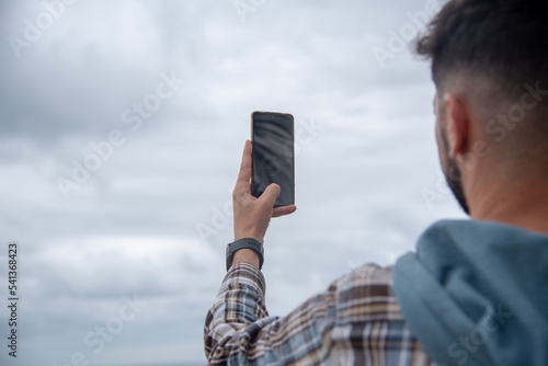 Un hombre moreno de espaldas sacando una foto al cielo con el teléfono móvil y una reloj inteligente con una camisa de cuadros.  photo