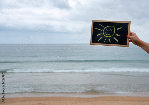 Mano de mujer con uñas rojas sujetando una pizarra negra con bordes de madera con un sol sonriente dibujado con tizas con una playa de fondo, el mar y las olas durante un día nublado de mal tiempo