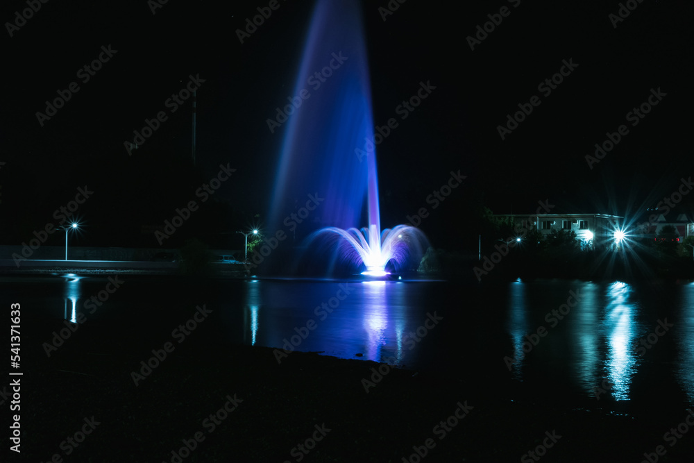 A beautiful high blue with purple fountain in the lake. A night fountain with a reflection in the water. Also in the water are reflected street lights.
