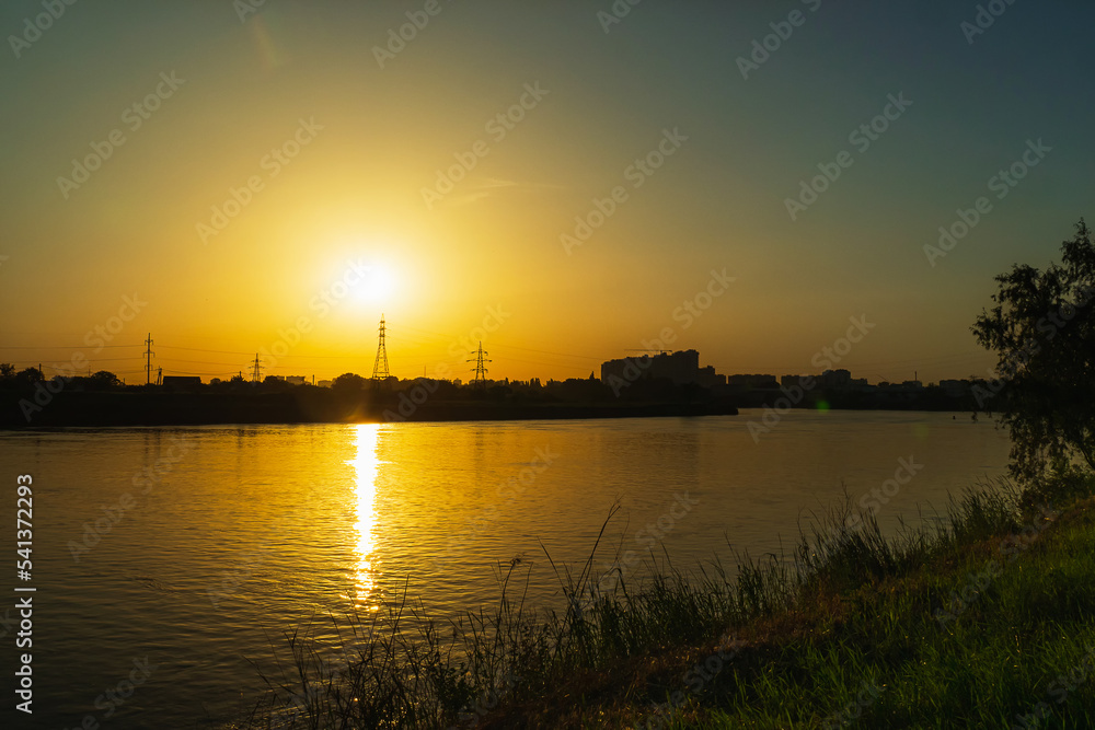 Sunset on the background of the river. The orange sun sets over the high-voltage power lines. In the foreground a bank of green grass. Sunset over the river in a small town.