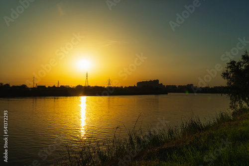 Sunset on the background of the river. The orange sun sets over the high-voltage power lines. In the foreground a bank of green grass. Sunset over the river in a small town.