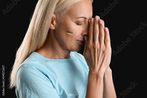 Mature woman with drawn flag of Ukraine praying on black background, closeup