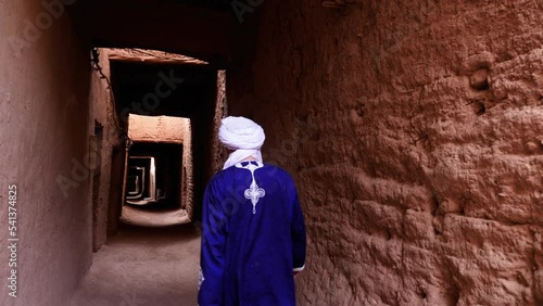 A traditionally dressed Bedouin Moroccan man wearing a blue gandoura and a turban walks inside the Kasbah in Mhamid, Morocco. Authentic rural scene of South Morocco. 4k. photo