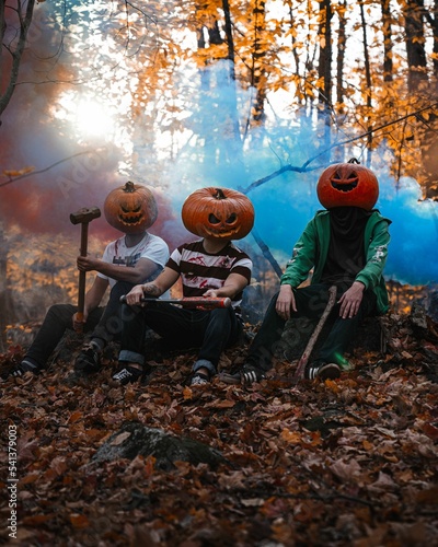 Three guys wearing pumpkins on their heads and sitting in a park during fall with smoke background