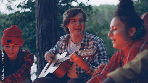 Handsome young man tourist is playing the guitar and smiling while his happy friends are singing and having fun resting around fire near lake or river. Green trees are visible. © silverkblack