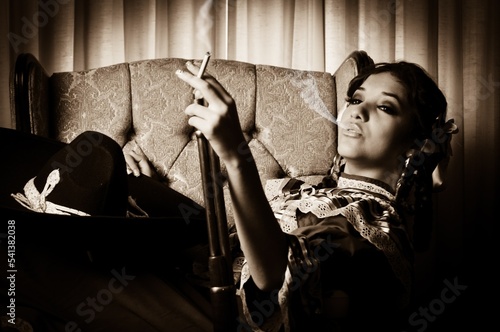 Retrato de una mujer modelo adolescente sentada mientras viste un traje típico de Adelita de la Revolución Mexicana, un sombrero de charro, un rifle, y fumando un cigarro photo