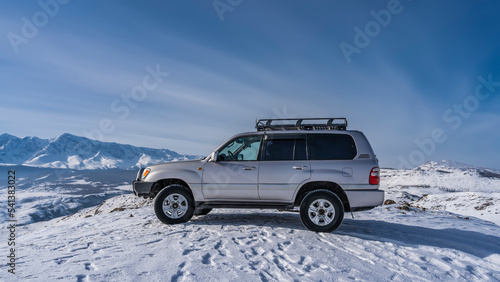 The SUV is standing on a snow-covered high-altitude plateau. Footprints are visible all around. A mountain range against a blue sky. Copy space. Altai