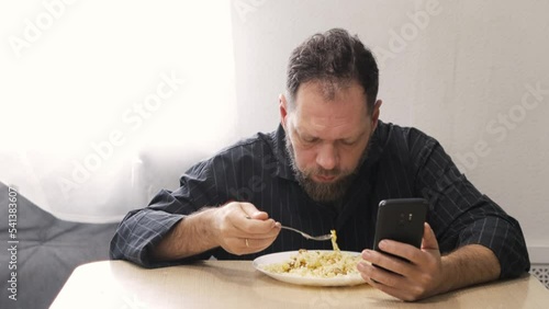 a tired and sleepy bearded man in pajamas eats pasta with meat and vegetables and watches the news on the smartphone screen