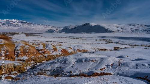 The bright red-orange hillsides are covered with snow. Dry grass and footprints in the valley. A mountain range against the blue sky. Altai Mars. Kyzyl Chin