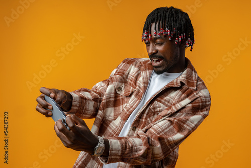 Young african man dressed in casual playing games on mobile phone over yellow background. © fotofabrika