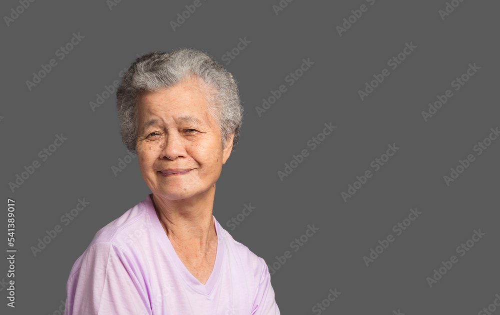 An elderly Asian woman looking at the camera with a smile while standing on a grey background An elderly Asian woman looking at the camera with a smile while standing on a grey background