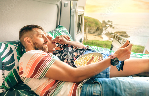 Young couple watching a movie on the tablet eating popcorn lying on bed of their camper van with doors open towards the coastal landscape