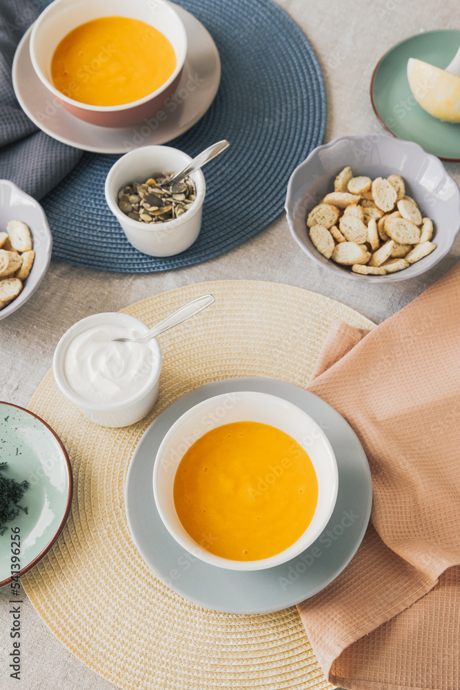 Top view of bowl with pumpkin soup. Near pumpkin seeds in a bowl, sour cream, crackers and second soup bowl. The table is covered with a linen tablecloth, with yellow and blue placemats. Top view of bowl with pumpkin soup. Near pumpkin seeds in a bowl, sour cream, crackers and second soup bowl. The table is covered with a linen tablecloth, with yellow and blue placemats.