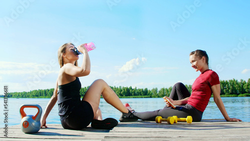 Two beautiful, athletic blonde girls, women have rest after training outdoors, drink water, talk, laugh. Lake, river, blue sky and forest in the background, summer sunny day. High quality photo © djtrenerfotolia