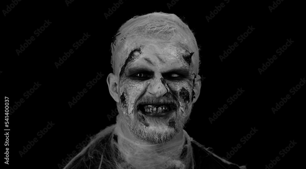 Fototapeta premium Portrait of sinister man in carnival costume of Halloween crazy zombie with bloody wounded scars face trying to scare isolated on black room. Horror theme of cosplay wounded undead, beast, monster