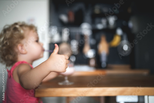 The child drinks a smoothie from a glass through a straw at home in the kitchen and shows the thumb of the hand, the gesture Cool and delicious. Healthy breakfast.