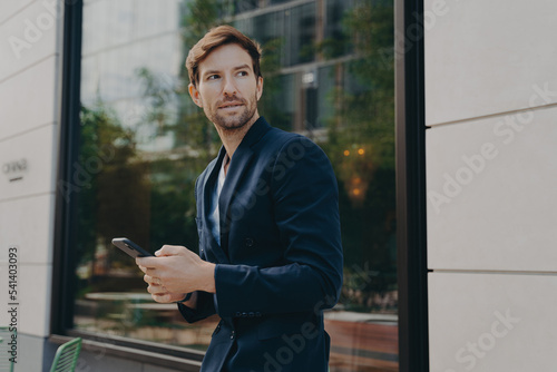 Businessman in dark suit holding smartphone and looking aside while walking city street