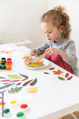A little girl makes crafts on an autumn theme. A three-year-old girl paints a sheet with yellow paint with her left hand. The girl got her fingers dirty. There is a white wall in the background.