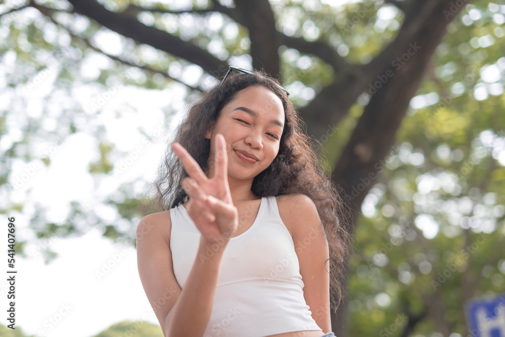 A low-angle shot of a beautiful young woman wearing a white tank top holds up a peace sign as she poses for the camera on a bright morning. A low-angle shot of a beautiful young woman wearing a white tank top holds up a peace sign as she poses for the camera on a bright morning.