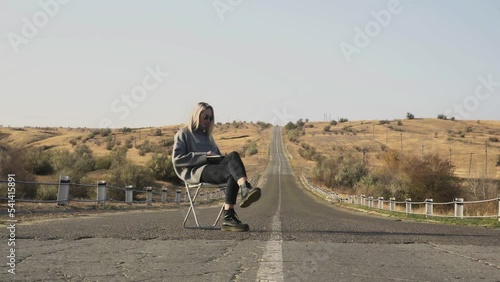 Young blonde woman in sunglasses with notebook sits on chair on country road and poses on camera photo