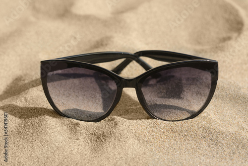 Stylish sunglasses with black frame on sandy beach, closeup