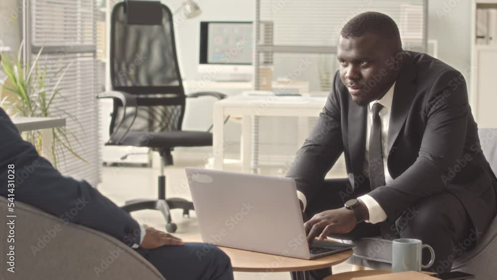 Two African American male business partners in suits having conversation and shaking hands during meeting in contemporary office Two African American male business partners in suits having conversation and shaking hands during meeting in contemporary office