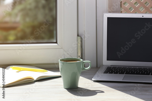 Cup of coffee, laptop with blank screen, stationery on wooden window sill. Space for text