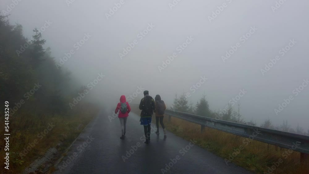 Group of friends are walking along a deserted road on a foggy day in the mountains. Travelers with backpacks in slow motion braving bad weather together in nature Group of friends are walking along a deserted road on a foggy day in the mountains. Travelers with backpacks in slow motion braving bad weather together in nature
