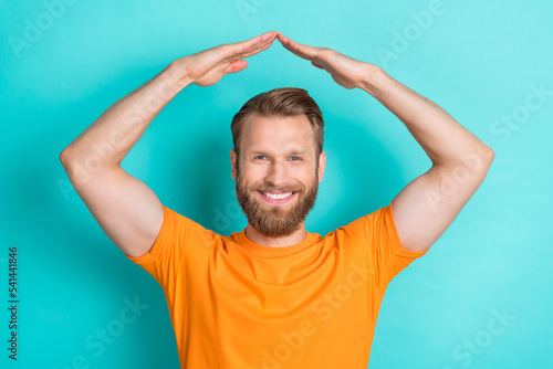 Portrait of cheerful positive guy with blond hairdo wear orange t-shirt hands showing roof over head isolated on teal color background