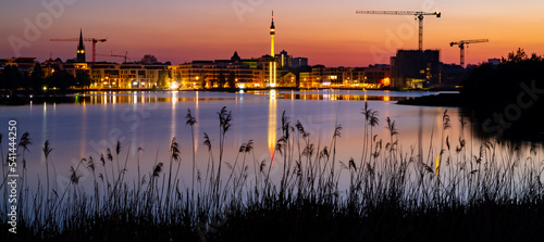Dortmund wide angle nighttime panorama. German city with downtown skyline, modern buildings and pond. Evening sky with clouds and reflection in Phoenix lake at blue hour with colorful illumination