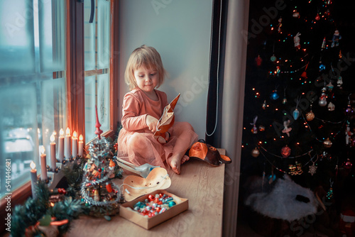 Cute little girl is dressing up a tiny Christmas tree and trying on New Year's animal masks. Image with selective focus and toning. 