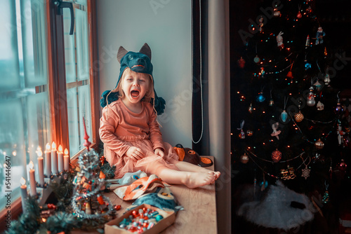 Cute little girl is dressing up a tiny Christmas tree and trying on New Year's animal masks. Image with selective focus and toning. 