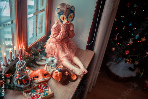 Cute little girl is dressing up a tiny Christmas tree and trying on New Year's animal masks. Image with selective focus and toning. 